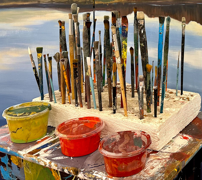 Foreground image featuring three paint containers: brown butter, pomegranate red, and crimson red, along with a paintbrush holder containing used brushes on a table covered in paint strokes. In the background, a painting depicts a serene body of water reflecting mountains.